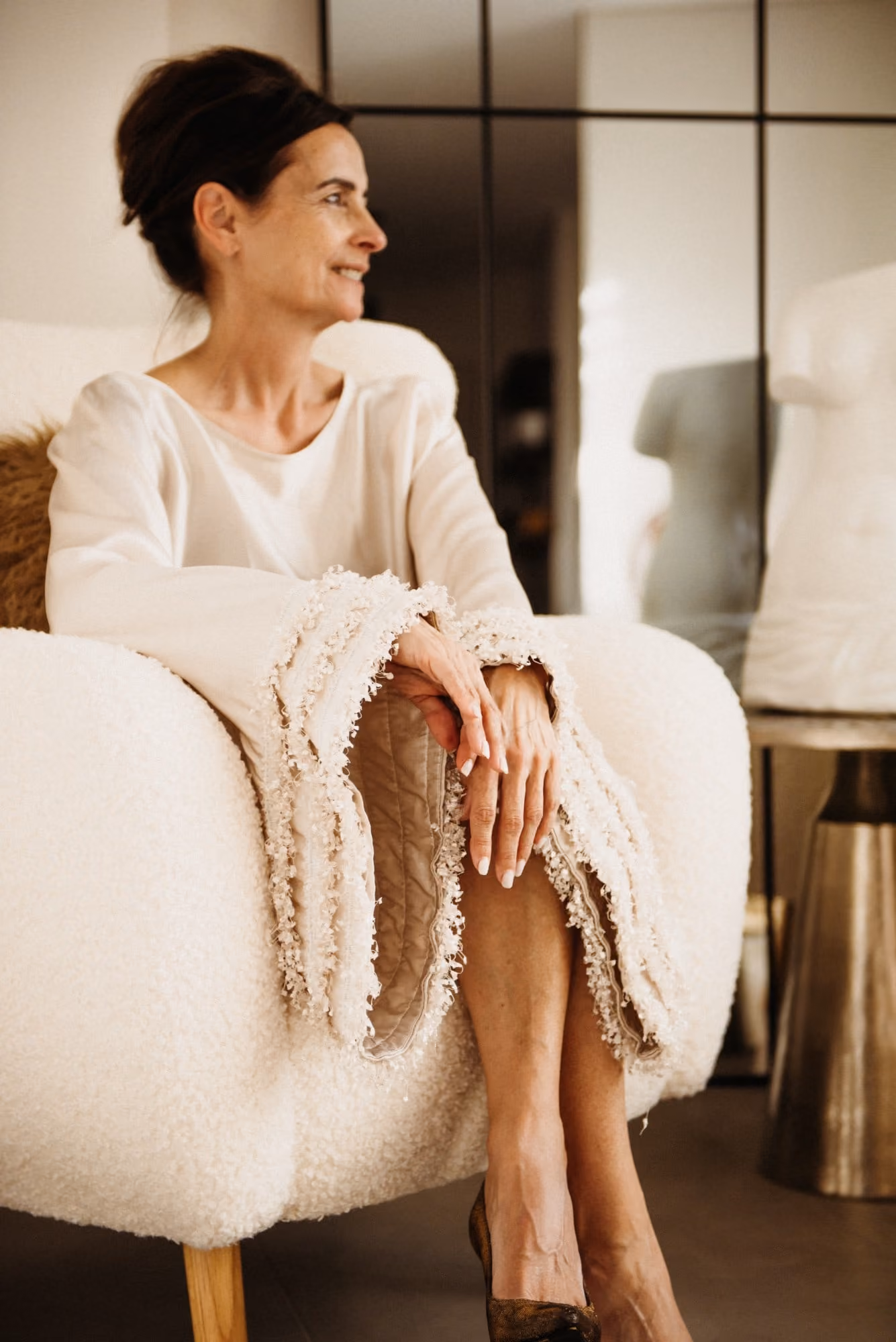Elle Wilson seated in a white armchair in a cream top with fringed sleeves, her hands resting on her crossed knee, smiling as she looks to one side